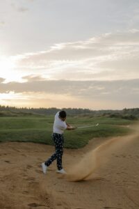A golfer swings in a sand trap at sunset on a scenic golf course.