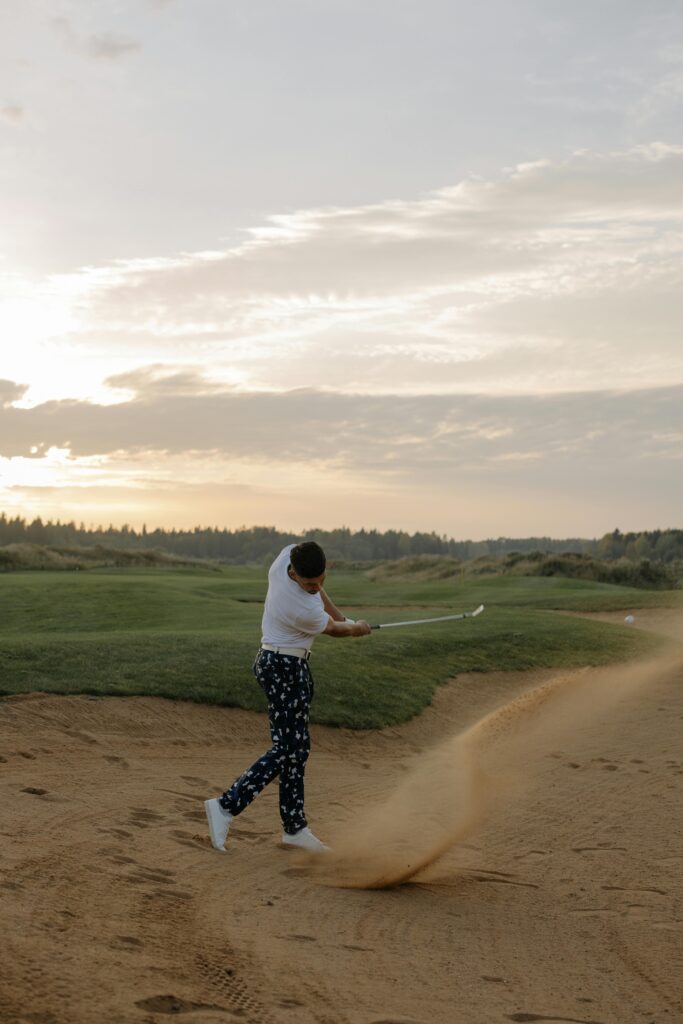 A golfer swings in a sand trap at sunset on a scenic golf course.